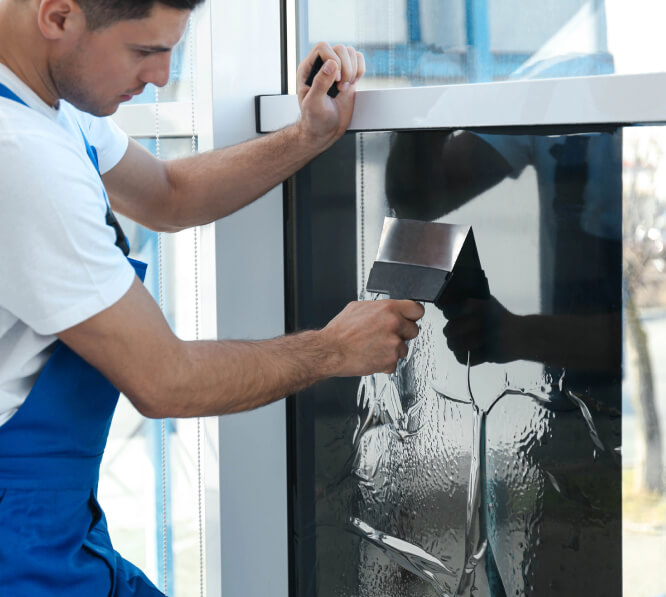 A person in blue overalls removes a dark window film from a glass pane using a metal scraper tool.