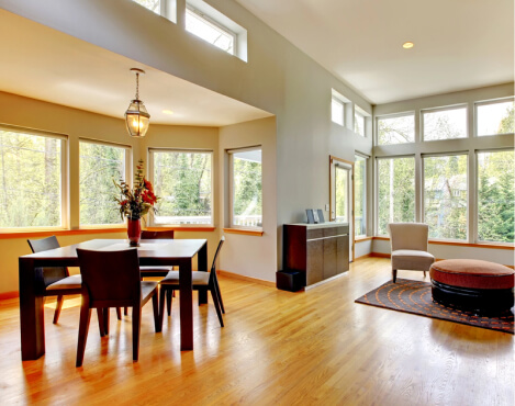 Modern dining and living area with wood floors, large windows, a dark dining set, an armchair, and a round ottoman. Natural light fills the space, highlighting the outdoor view.