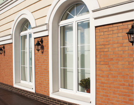 Two arched windows with white frames on a brick and beige exterior wall. Black wall lanterns are mounted beside each window, and a potted plant is visible through one window.