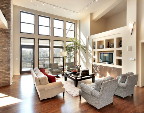 Modern living room with large windows, neutral-colored sofas and chairs, a coffee table, a tall plant, built-in shelves, a TV, and wooden flooring.