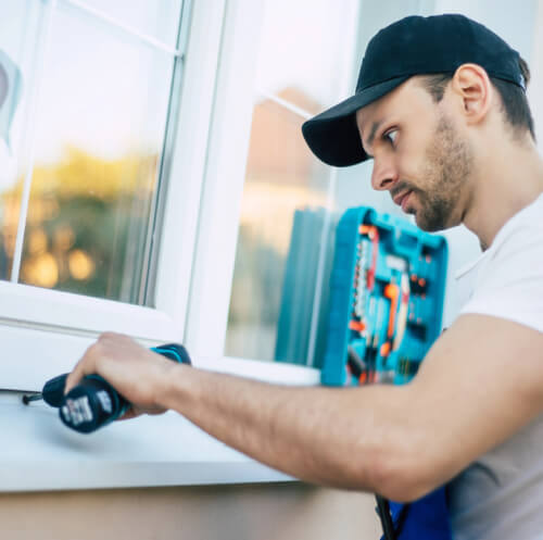 Man in a black cap uses a power drill to install or repair a white window frame, with a toolbox in the background.